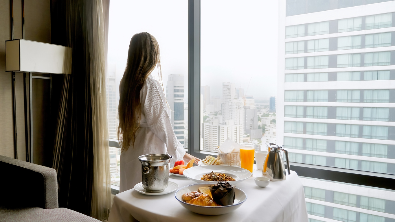Eine Frau mit braunen langen Haaren und weißem Bademantel steht an einem riesigen Fenster und blickt auf eine Skyline mit Wolkenkratzern. Neben ihr steht ein gedeckter Frühstückstisch. Eine Frau mit braunen langen Haaren und weißem Bademantel steht an einem riesigen Fenster und blickt auf eine Skyline mit Wolkenkratzern. Neben ihr steht ein gedeckter Frühstückstisch.
